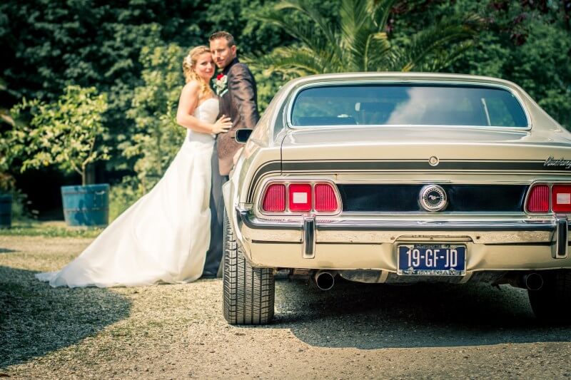 Ford Mustang Trouwauto Zelf Rijden (Klassieke Verhuur) Bruidspaar poseert met een vintage Ford Mustang in een tuin